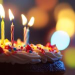 A close-up, low-angle shot of a chocolate birthday cake topped with white frosting and colorful sprinkles. Five thin, lit candles with flickering yellow flames stand atop the cake. The background is a soft, warm bokeh of golden, yellow, and blue lights, creating a festive and celebratory atmosphere.