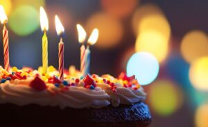 A close-up, low-angle shot of a chocolate birthday cake topped with white frosting and colorful sprinkles. Five thin, lit candles with flickering yellow flames stand atop the cake. The background is a soft, warm bokeh of golden, yellow, and blue lights, creating a festive and celebratory atmosphere.
