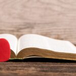 An open Bible rests on a rustic wooden surface, with a small, vibrant red heart-shaped ornament leaning against its left page. The background is a soft-focus, light-colored wooden wall, creating a warm and peaceful atmosphere.