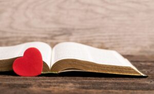 An open Bible rests on a rustic wooden surface, with a small, vibrant red heart-shaped ornament leaning against its left page. The background is a soft-focus, light-colored wooden wall, creating a warm and peaceful atmosphere.