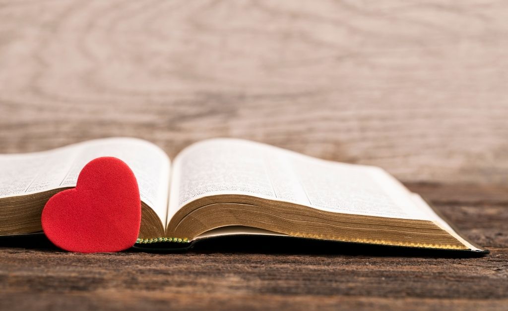 An open Bible rests on a rustic wooden surface, with a small, vibrant red heart-shaped ornament leaning against its left page. The background is a soft-focus, light-colored wooden wall, creating a warm and peaceful atmosphere.