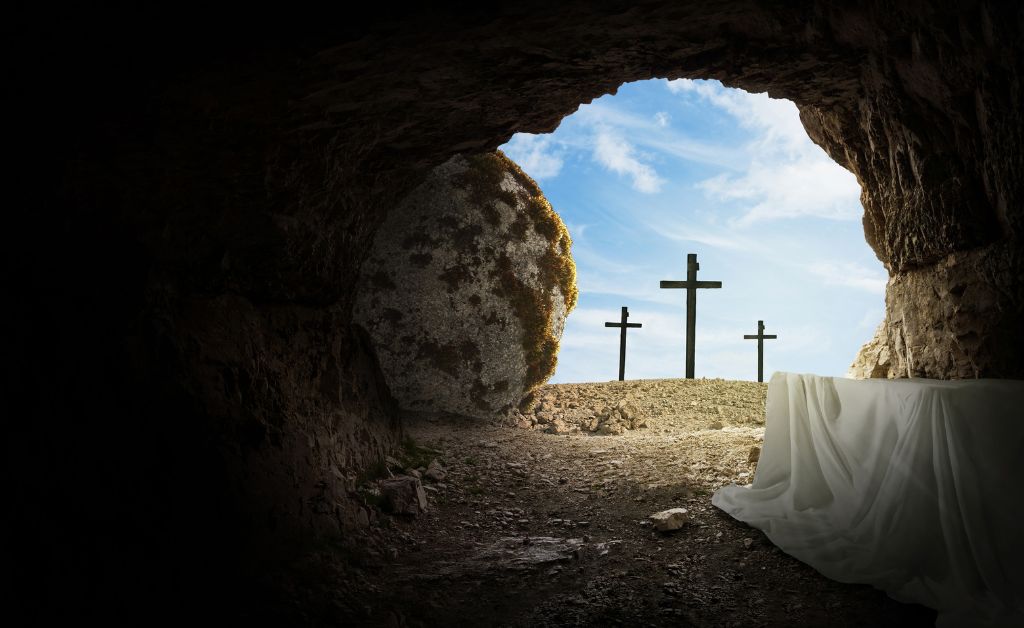 An empty stone tomb viewed from the inside looking out. In the foreground, a white burial cloth lies draped on a stone bench. The large circular stone that sealed the entrance has been rolled aside, revealing a bright blue sky with soft white clouds. In the distance, three wooden crosses stand silhouetted on a hill against the horizon.