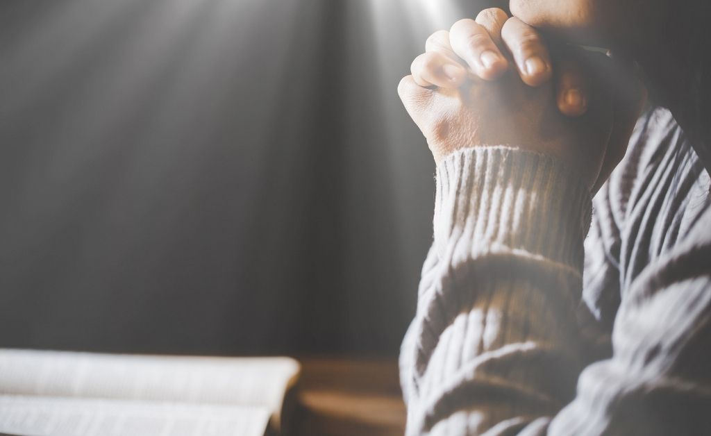 A close-up, side profile of a person’s hands clasped together in prayer over an open book, likely a Bible. The person is wearing a grey ribbed sweater, and their head is slightly bowed. Soft, dramatic rays of light shine down from the upper left corner against a dark, blurred background, creating a peaceful and spiritual atmosphere.