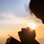 A silhouette of a woman with her head bowed and hands clasped in prayer against a warm, glowing sunset. She holds a small book, likely a Bible, as the sun's light peeks through her fingers.
