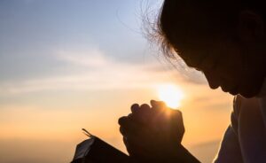 A silhouette of a woman with her head bowed and hands clasped in prayer against a warm, glowing sunset. She holds a small book, likely a Bible, as the sun's light peeks through her fingers.