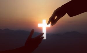 A silhouette of two hands reaching toward each other against a sunset backdrop. A glowing white cross is positioned in the center, between the hands, over a range of distant mountains.