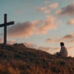 A man sits in quiet prayer on a grassy hillside at sunset, facing a simple wooden cross. The soft orange and pink hues of the evening sky create a serene, spiritual atmosphere, emphasizing a moment of deep reflection, faith, and peace in nature.