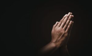 : A close-up of two hands pressed together in a prayer position against a dark, moody background. The lighting emphasizes the skin texture and the solemn gesture.