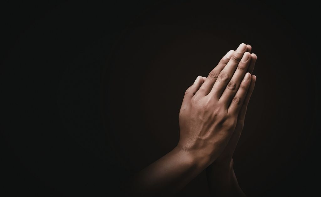 : A close-up of two hands pressed together in a prayer position against a dark, moody background. The lighting emphasizes the skin texture and the solemn gesture.