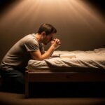 A man in a gray t-shirt and dark pants kneels on a carpeted floor beside a wooden bed, his hands clasped in prayer. An open book, resembling a Bible, rests on the white bedsheets in front of him. A dramatic, soft light shines down from above, illuminating him and the bed while leaving the rest of the bedroom in deep shadow.